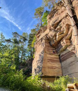Person klettert an einer hohen, gestreiften Felswand im Wald unter blauem Himmel.