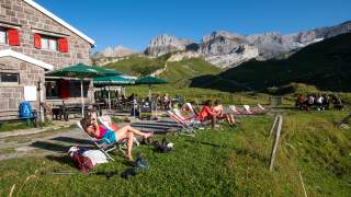 Menschen entspannen auf Liegestühlen vor einer Berghütte in einer sonnigen Alpenlandschaft mit grünen Wiesen und felsigen Bergen im Hintergrund.
