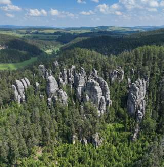 Luftaufnahme von markanten Sandsteinfelsen, die aus einem dichten Nadelwald herausragen, im Hintergrund grüne Hügel und blauer Himmel mit Wolken.