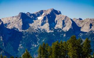 Hohe, felsige Berge mit spärlichen Schneefeldern im Sonnenlicht, im Vordergrund grüne Nadelbäume, blauer Himmel im Hintergrund.