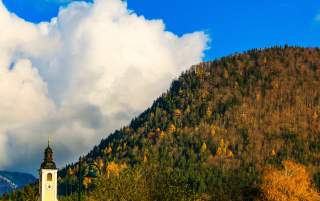 Kirchturm mit Uhr vor bewaldetem Berghang im Herbst, darüber blauer Himmel mit großen weißen Wolken.
