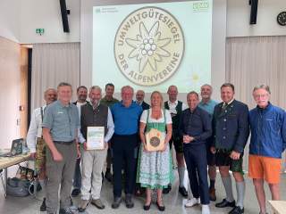 Eine Gruppe von 13 Personen steht in einem Raum vor einer Leinwand mit dem Logo „Umweltgütesiegel der Alpenvereine“, zwei Personen halten Urkunden und eine Frau trägt ein Dirndl.