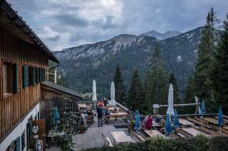Außenterrasse einer Berghütte mit mehreren Holztischen und Sitzbänken, umgeben von Nadelbäumen und Bergen im Hintergrund unter bewölktem Himmel.