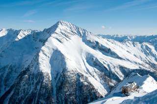Schneebedeckte Berge unter klarem blauem Himmel mit einer Berghütte im Vordergrund auf einer verschneiten Fläche.