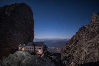 Berghütte mit beleuchteten Fenstern liegt zwischen zwei Felswänden unter einem klaren Sternenhimmel, im Hintergrund sind entfernte Berge und Täler zu sehen.