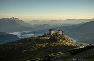 Berghütte auf einem grasbewachsenen Gipfel mit dahinterliegenden, in Nebel gehüllten Bergketten im Morgenlicht.