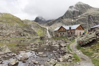 Bergsee mit Felsen und einem steinernen Gebäude am Ufer, im Hintergrund ein Wasserfall und bewölkte Berge. Ein schmaler Wanderweg führt zum Haus.