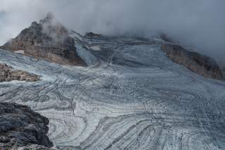 Verschneites, bergiges Landschaftsbild mit sichtbaren Gletscherspalten und Wolken, die die Bergspitzen teilweise verdecken.