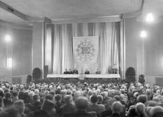 Schwarzweißfoto einer großen Versammlung in einem Saal mit einem Podium, auf dem fünf Männer sitzen, und einem Redner am Pult; im Hintergrund ein Banner mit der Aufschrift „Deutscher Alpenverein“.