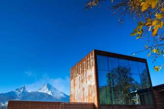 Modernes Gebäude mit spiegelnden Glasfenstern und rostbraunen Wänden, auf denen „Haus der Berge“ steht, vor einer Berglandschaft mit schneebedeckten Gipfeln und klarem blauem Himmel.