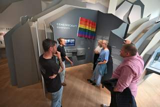 Fünf Personen stehen in einem Ausstellungsbereich mit der Überschrift „Gemeinschaft“ und einer Regenbogenflagge an der Wand, während sie sich unterhalten.