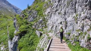 Eine Person mit Wanderausrüstung geht auf einem schmalen Steg entlang einer felsigen Bergwand, umgeben von Grünflächen und unter klarem, blauem Himmel.