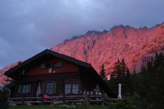 Berghütte im Vordergrund, dahinter ein von der Abendsonne rot angestrahltes Felsmassiv, teils bewaldet, wolkiger Himmel.