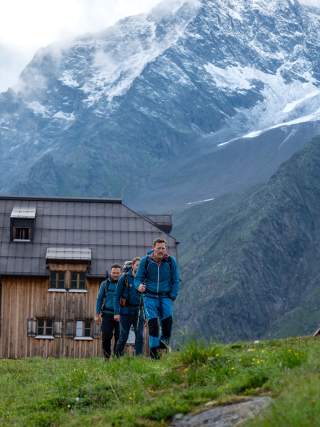 Drei Wanderer in Outdoor-Kleidung gehen vor einer Berghütte mit schneebedeckten Bergen im Hintergrund.