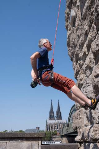 Ein älterer Mann mit Klettergurt hängt an einer Kletterwand, im Hintergrund sind der Kölner Dom und die Hohenzollernbrücke zu sehen.