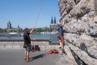 Zwei Männer klettern an einer steinernen Mauer am Rheinufer; im Hintergrund sind der Kölner Dom, die Altstadt und vorbeifahrende Schiffe zu sehen.