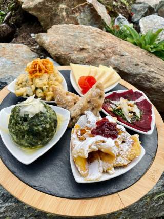 Runde Platte mit verschiedenen kleinen Gerichten, darunter Käsescheiben, Spinatknödel, Rote-Bete-Scheiben, Spätzle mit Röstzwiebeln, Gebäck und ein Dessert mit Puderzucker und Fruchtkompott. Im Hintergrund sind Felsen und Pflanzen zu sehen.