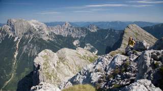 Ein Wanderer mit Helm und Rucksack klettert auf felsigem Gebirgspass mit Aussicht auf hohe Berge und ein Tal unter blauem Himmel.