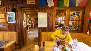 In einer gemütlichen Holzhütte sitzt eine Person in gelber Jacke an einem Tisch und isst, während im Hintergrund weitere Gäste an Tischen sitzen. Die Wände sind mit Karten, Hinweisschildern und bunten Gebetsfahnen dekoriert.
