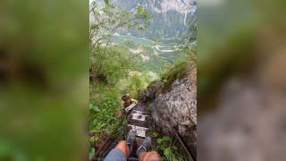 Blick von oben auf eine steile Metalltreppe im Gebirge, auf der eine Person hinaufklettert, umgeben von grüner Vegetation und mit Blick ins Tal auf einen Fluss und bewaldete Berge.