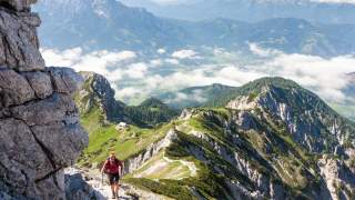 Wanderer erklimmt felsigen Bergpfad mit weiter Aussicht auf grüne Berge und Wolken darunter.