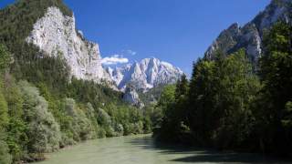 Fluss fließt durch bewaldetes Tal mit steilen Felsklippen und schneebedeckten Bergen im Hintergrund unter klarem blauem Himmel.