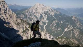 Ein Wanderer mit Rucksack steht auf einem grasbewachsenen Felsvorsprung und blickt auf eine bergige Landschaft mit schroffen Felsen und Tälern unter klarem Himmel.