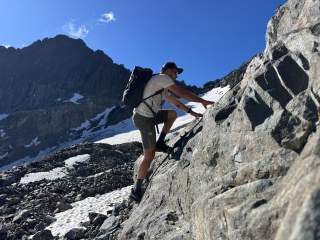 Ein Mann mit Rucksack klettert bei sonnigem Wetter an einer felsigen Bergwand, im Hintergrund sind schneebedeckte Berge.