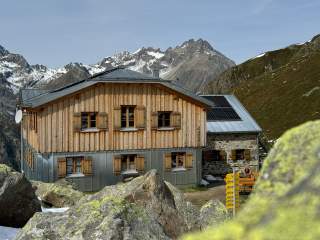 Berghütte aus Holz und Stein mit Solarpanel auf dem Dach, umgeben von Felsen und schneebedeckten Bergen im Hintergrund.