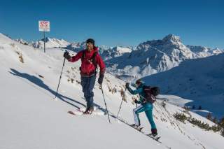 Zwei Skitourengeher steigen auf einer schneebedeckten Bergflanke bei klarem Himmel auf, neben ihnen ein Schild mit der Aufschrift „Stopp!“. Im Hintergrund sind schneebedeckte Berggipfel zu sehen.