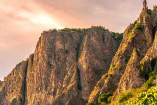 Felsige Bergspitzen im warmen Licht der Abendsonne mit vereinzeltem Grün auf den Gipfeln und am Hang. Am oberen linken Felsen ist eine Mauer oder Festung zu erkennen.