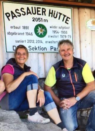 Zwei lächelnde Personen sitzen vor einer Holzwand mit einem Schild, auf dem 