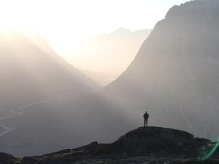 Person steht auf einem Felsen und blickt auf ein nebliges Tal zwischen hohen Bergen im Morgenlicht.