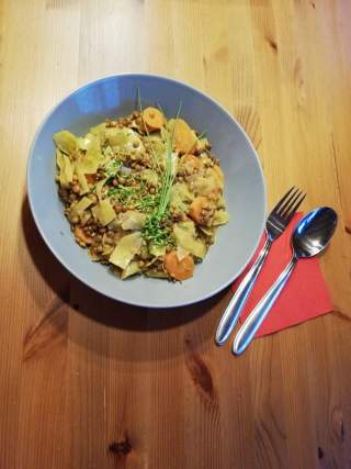 Blaue Schale mit einem vegetarischen Gericht aus Linsen, Karottenscheiben und Kräutern, daneben liegen eine Gabel und ein Löffel auf einer roten Serviette auf einem Holztisch.