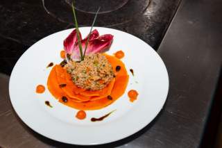 Weiße Tellerpräsentation mit einer Kugel Gemüsereis auf dünnen, runden Süßkartoffelscheiben, garniert mit Radicchio, Schnittlauch, Kürbiskernen und kleinen Tupfen roter Soße.