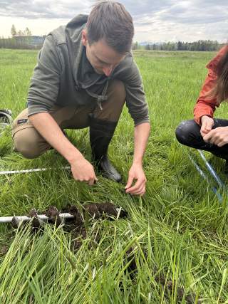 Zwei Personen sammeln Proben auf einer Moorfläche; eine Person hält frisch ausgegrabene Erde und Gras, während die andere daneben hockt. Im Vordergrund liegt eine Erdsonde mit entnommenem Boden.