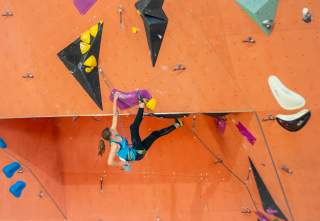 Eine Frau klettert an einer orangefarbenen Kletterwand in einer Halle, sie trägt Kletterschuhe und ist mit einem Seil gesichert. Verschiedene bunte Griffe und Volumen sind an der Wand befestigt.