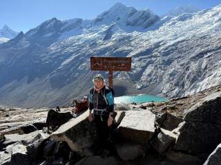 Person mit Wanderausrüstung steht vor Gipfelschild „Punta Union 4750 m.s.n.m.“ in einer felsigen, bergigen Landschaft mit schneebedeckten Gipfeln und einem türkisfarbenen Bergsee im Hintergrund.