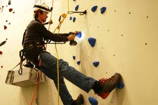 Ein Mann mit Schutzhelm und Kletterausrüstung befestigt blaue Griffsteine an einer Kletterwand mit einem Akkuschrauber.