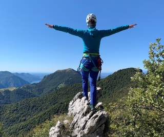 Eine Person mit Kletterausrüstung und Helm steht auf einem Felsen, breitet die Arme aus und blickt auf bewaldete Berge unter blauem Himmel.