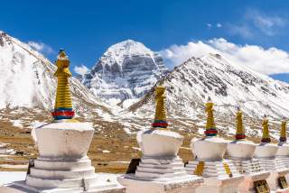 Schneebedeckte Berge und mehrere tibetische Stupas im Vordergrund unter blauem Himmel.