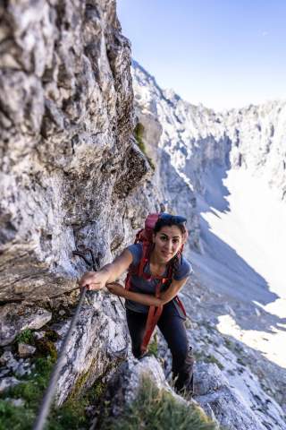 Frau mit Rucksack klettert an einer Felswand und hält sich dabei an einem Sicherungsseil fest, im Hintergrund eine felsige Berglandschaft.