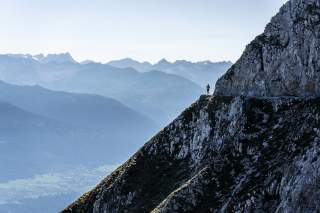 Ein einzelner Wanderer steht auf einem schmalen Bergpfad an einer steilen Felswand, im Hintergrund sind mehrere Bergketten in blauem Dunst zu sehen.