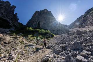 Zwei Wanderer auf einem steinigen Gebirgspfad bei sonnigem Wetter mit einer markanten Felswand im Hintergrund. Die Sonne scheint am klaren blauen Himmel und erzeugt einen Sterneneffekt.