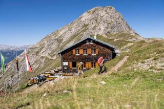 Bergwanderer steigen auf einem Pfad neben einer Berghütte mit Außenterrasse, im Hintergrund ragen steile Felswände und ein klarer blauer Himmel empor.