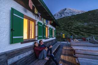 Zwei Personen sitzen abends entspannt auf einer Holzbank vor einer Berghütte mit grünen Fensterläden, im Hintergrund sind Bergwiesen und ein Felsgipfel zu sehen.
