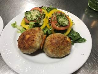 Weißer Teller mit zwei gebratenen Frikadellen und einem frischen Salat aus gelber Paprika, Tomatenscheiben, Gurkenscheiben und Blattsalat, garniert mit Kresse.
