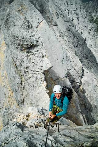 Ein Bergsteiger mit Helm und Kletterausrüstung steigt eine steile Felswand über Eisenbügel empor. Die Umgebung besteht aus grauem, schroffem Fels.