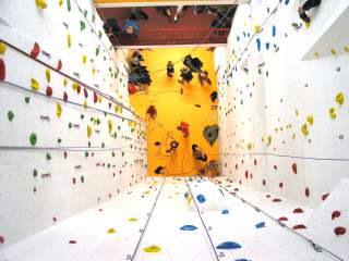 Blick von oben auf eine helle Indoor-Kletterwand mit bunten Griffen; unten stehen mehrere Menschen auf gelbem Boden.