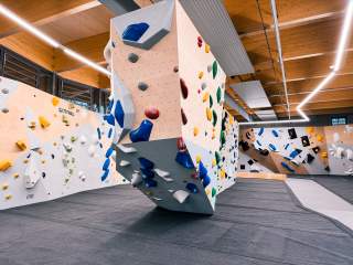 Moderner, heller Indoor-Boulderraum mit verschiedenen Kletterwänden und bunten Griffen sowie dick gepolstertem Boden. Decke und Wände sind mit Holz verkleidet, große Fenster sorgen für Tageslicht.